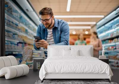 Happy man going through checklist on smart phone while shopping in supermarket. Wall mural