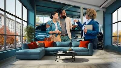 Happy multiracial couple receiving keys of their new car from saleswoman in showroom. Wall mural