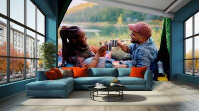 Happy multiracial couple toasting while relaxing in tent on camping. Wall mural