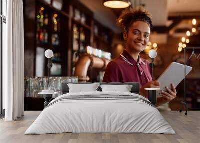 Happy waitress working on touchpad in  bar and looking at camera. Wall mural