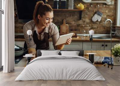 Happy woman reading recipe on touchpad while preparing food in the kitchen. Wall mural