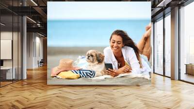 Happy woman texting on cell phone while relaxing with her dog on beach. Wall mural