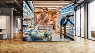 Happy woman using reminder on smart phone while shopping in supermarket. Wall mural