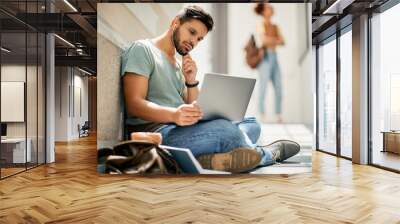 Male student reads e-mail on laptop while sitting on floor at university hallway. Wall mural