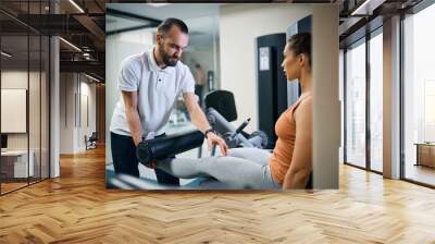 Physical therapist assists young woman with exercises on leg extension machine. Wall mural