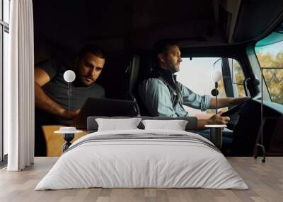 Professional driver driving truck while his colleague is using laptop in cabin. Wall mural