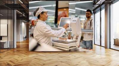 Smiling delicatessen section worker using grocery scale while serving her customer at supermarket. Wall mural