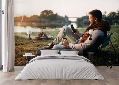 Smiling man using smart phone while relaxing by campfire in nature at sunset. Wall mural