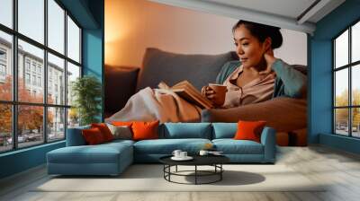 Young Asian woman dinks tea while reading book on the sofa in the evening. Wall mural