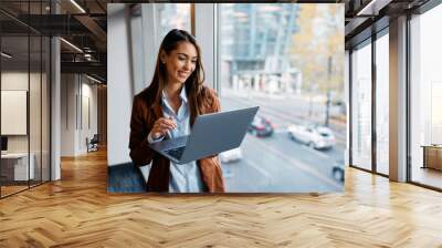 Young happy businesswoman working on laptop by  window in office. Wall mural