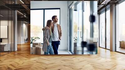 Young happy couple enjoys the view through window at their new home. Wall mural