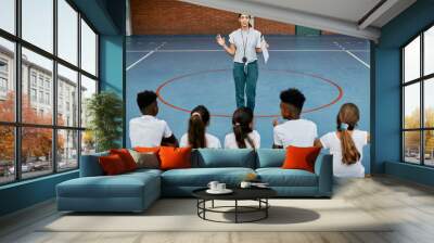 Young sports teacher communicates with group of school kids during physical education class. Wall mural