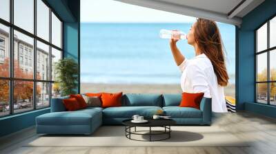 Young woman drinks water from bottle during summer day on beach. Wall mural