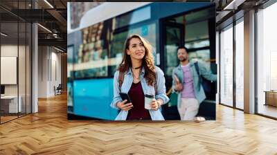 Young woman with takeaway coffee and cell phone at bus station. Wall mural