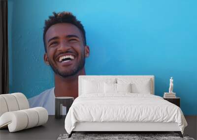 Capture a young Brazilian man in casual attire, standing against a bright blue background, laughing heartily with a genuine and infectious smile. Wall mural