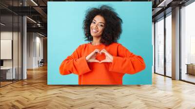 Adorable american woman in colorful shirt looking on camera and gesturing heart shape with hands, isolated over blue background Wall mural