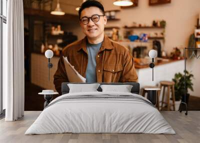 Adult asian man smiling and holding paper documents in cafe indoors Wall mural