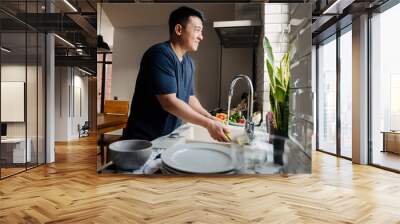 Adult brunette asian man wearing t-shirt washing dishes at kitchen Wall mural