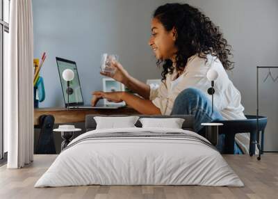 African american young woman drinking water and using laptop by desk Wall mural