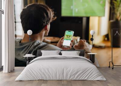 Back view of man watching football match and making bets at bookmaker's website in front of TV screen Wall mural