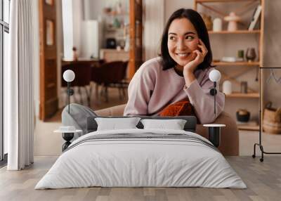 Beautiful asian woman smiling and looking away while sitting at home Wall mural