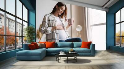 Beautiful happy pregnant woman smiling and drinking coffee while sitting on couch Wall mural