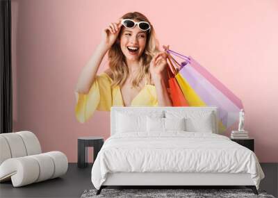 Beautiful happy young blonde woman posing isolated over pink wall background holding shopping bags. Wall mural