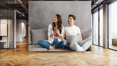 Beautiful man and woman sitting together on couch in gray interior, and looking aside on copyspace while using laptop and smartphone Wall mural