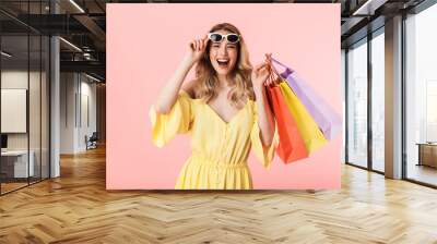 Beautiful shocked young blonde woman posing isolated over pink wall background holding shopping bags. Wall mural