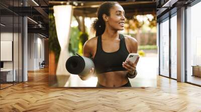 Black woman smiling and using cellphone while standing with yoga mat Wall mural