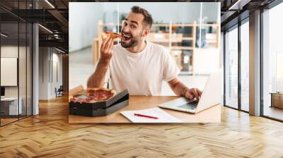 Caucasian joyful man smiling and eating pizza while working with laptop Wall mural