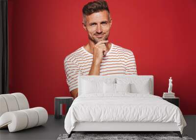 Caucasian man in striped t-shirt thinking and touching chin, isolated over red background Wall mural