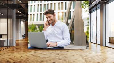 Cheerful happy young businessman sitting outdoors on the street on bench using laptop computer talking by mobile phone. Wall mural