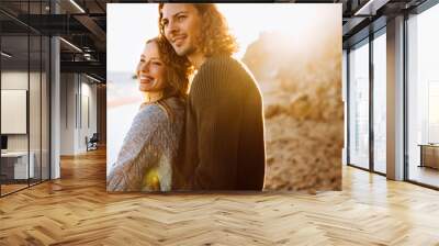 Cheerful lovely couple sweaters hugging while standing by the rocks at sunny beach Wall mural