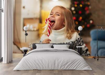 Close-up photo of funny little girl licks candy cane, looking at camera Wall mural