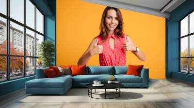 Close-up portrait of charming smiling brunette girl in red dress showing two thumbs up, looking at camera Wall mural