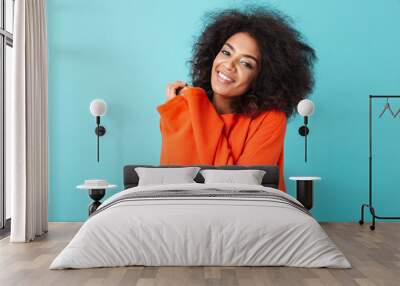 Colorful portrait of african american woman in red shirt with afro hairstyle looking on camera with smile pressing her arms to chest, isolated over blue background Wall mural