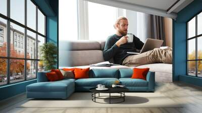 Concentrated young bearded man sitting in home using laptop computer. Wall mural