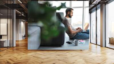 Concentrated young man using laptop computer sit on floor. Wall mural