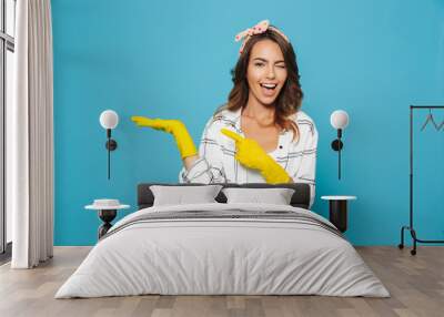 Content happy woman 20s holding copyspace on her palm while wearing yellow rubber gloves for hands protection during cleaning, isolated over blue background Wall mural