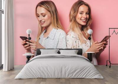 Cute cheerful beautiful blondes women friends posing isolated over pink wall background using mobile phones. Wall mural