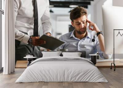 Displeased man sitting in office working with computer near his colleague who showing clipboard with documents. Wall mural