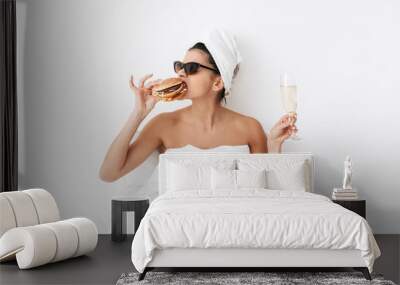 Emotional woman with towel on head lies in bed under blanket isolated over white wall background wearing sunglasses eat burger. Wall mural