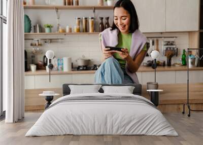 Excited asian woman playing online game on mobile phone while sitting in kitchen Wall mural