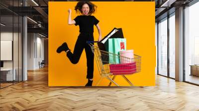 Excited woman making winner gesture while posing with shopping cart Wall mural