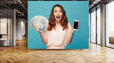 Excited young lady showing display of mobile phone holding money. Wall mural