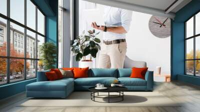 Full length photo of attractive modern man wearing eyeglasses looking at camera, while standing and working on laptop in office Wall mural