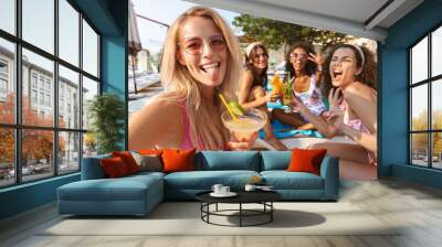 Group of women friends sitting outdoors in pool dressed in swimwears take a selfie by camera. Wall mural