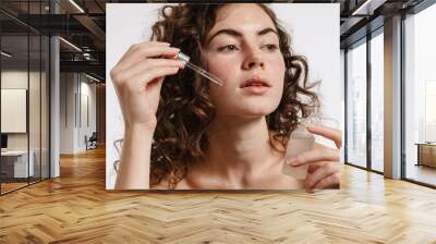 Half-naked curly woman applying face serum while looking at camera Wall mural