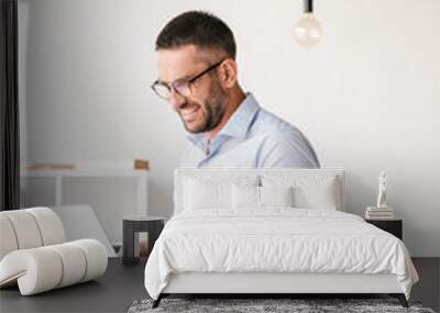 Handsome joyful man wearing white shirt sitting on table in office, and having business chat on silver laptop Wall mural